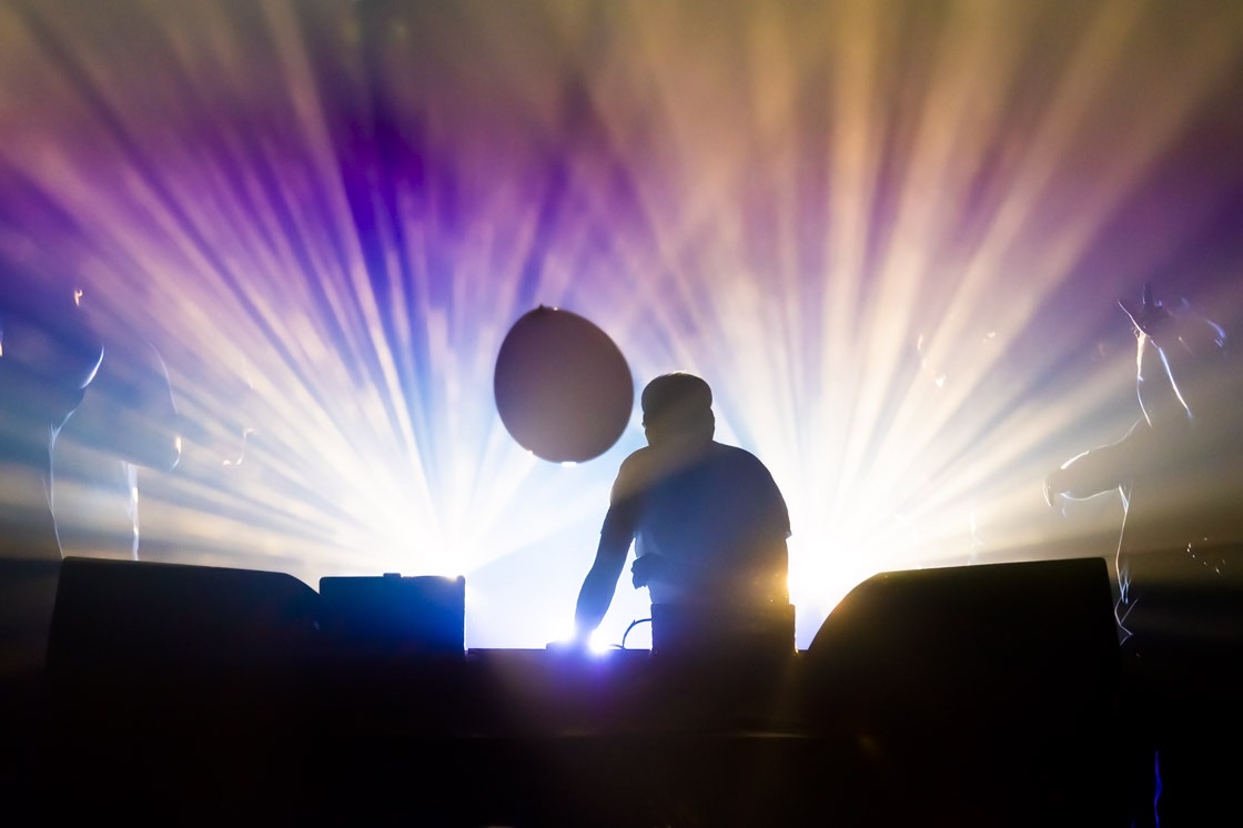 A DJ in silhouette leans over his laptop as purple and gold light beams fan out behind him and a balloon hangs in the air.