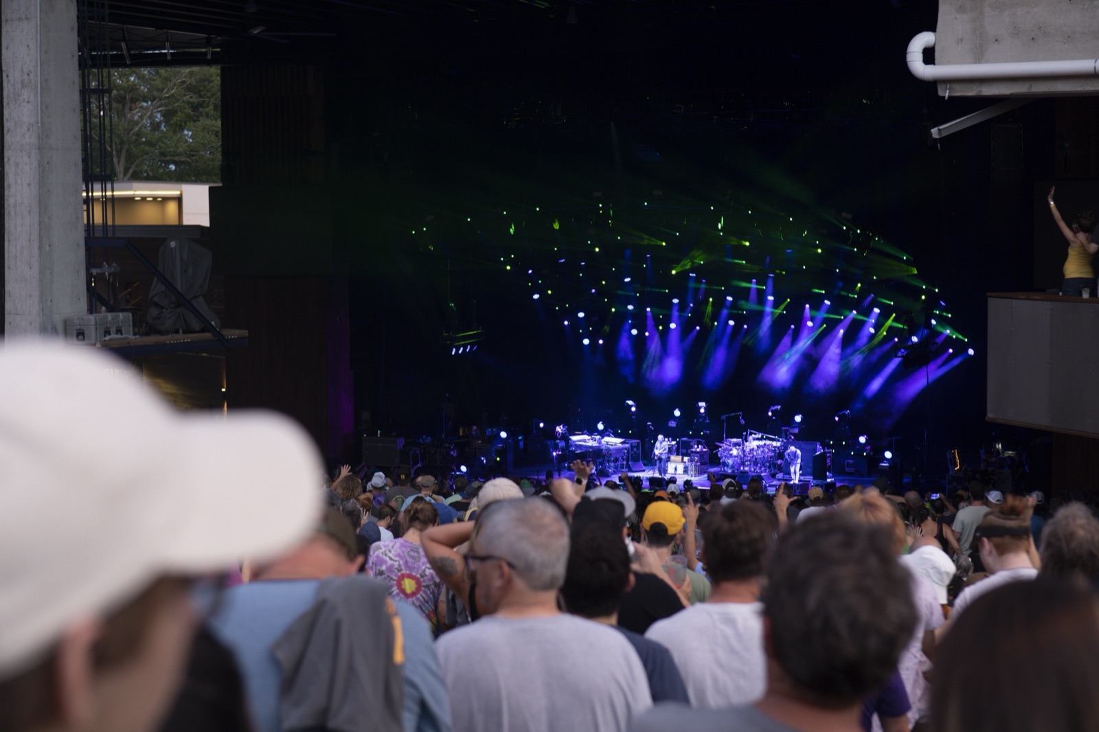 The band plays under fans of blue and green stage lights at the back of a covered pavilion, seen over the heads of the crowd.