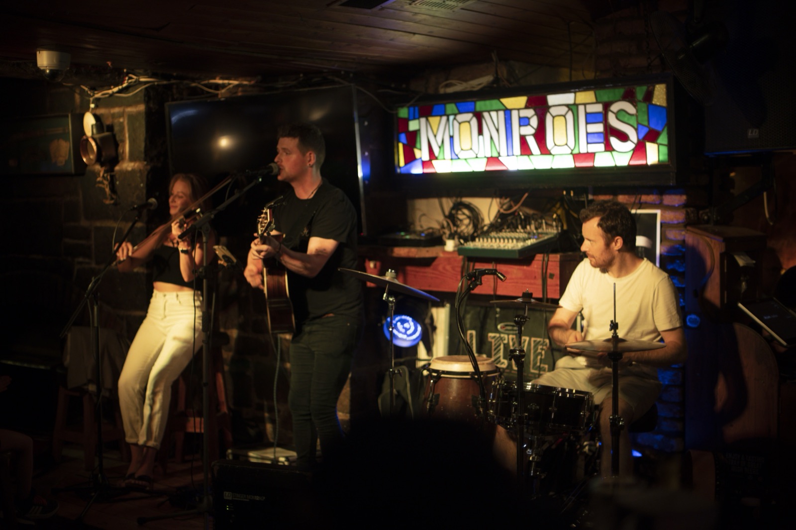 A fiddler, a guitarist and a drummer play on a small pub stage under a stained glass Monroe's sign.