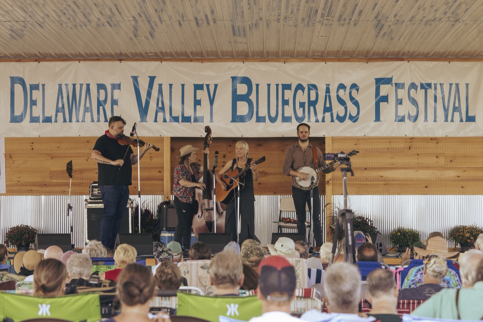 A four-piece bluegrass band with fiddle, upright bass, guitar and banjo plays under a Delaware Valley Bluegrass Festival banner to a seated crowd.