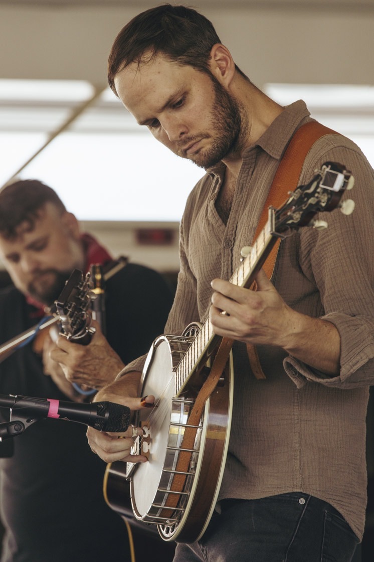 A banjo player looks down at his strings while a fiddler plays behind him at a festival stage mic.