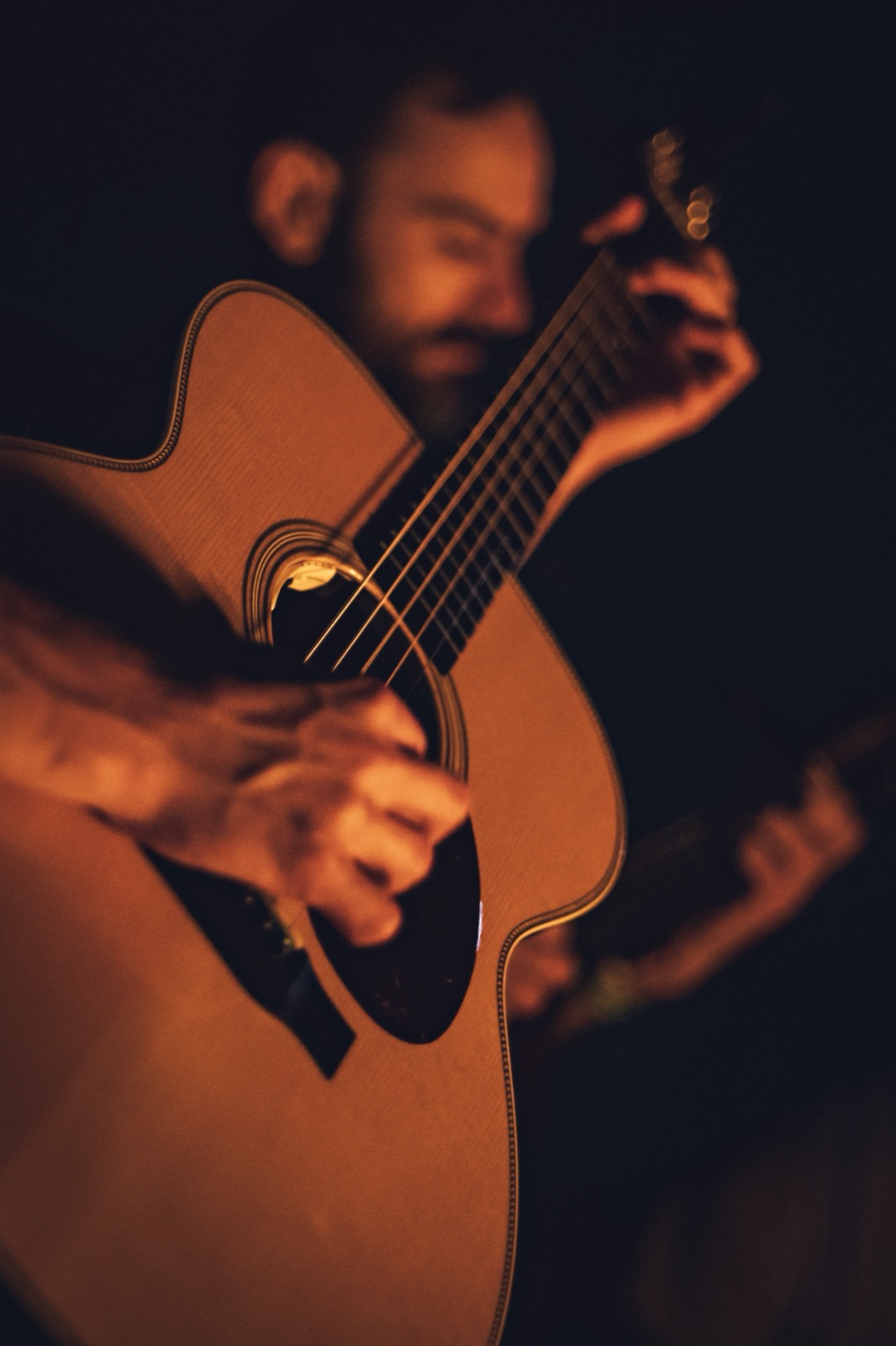 A bearded guitarist's hands work an acoustic guitar, lit orange against the dark.