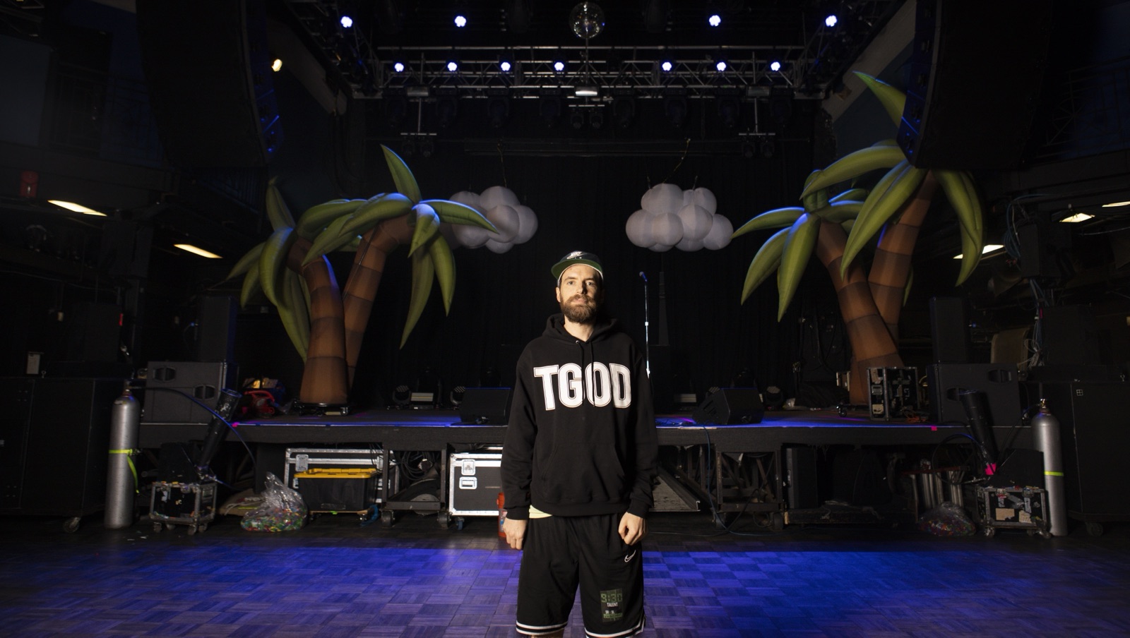 Girl Talk, in a black hoodie, stands alone on the empty stage before the show, between two inflatable palm trees.