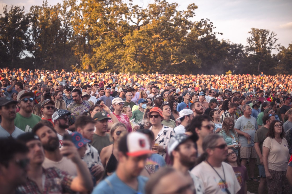 A packed lawn crowd looks toward the stage in warm late sun, with oak trees glowing behind them.