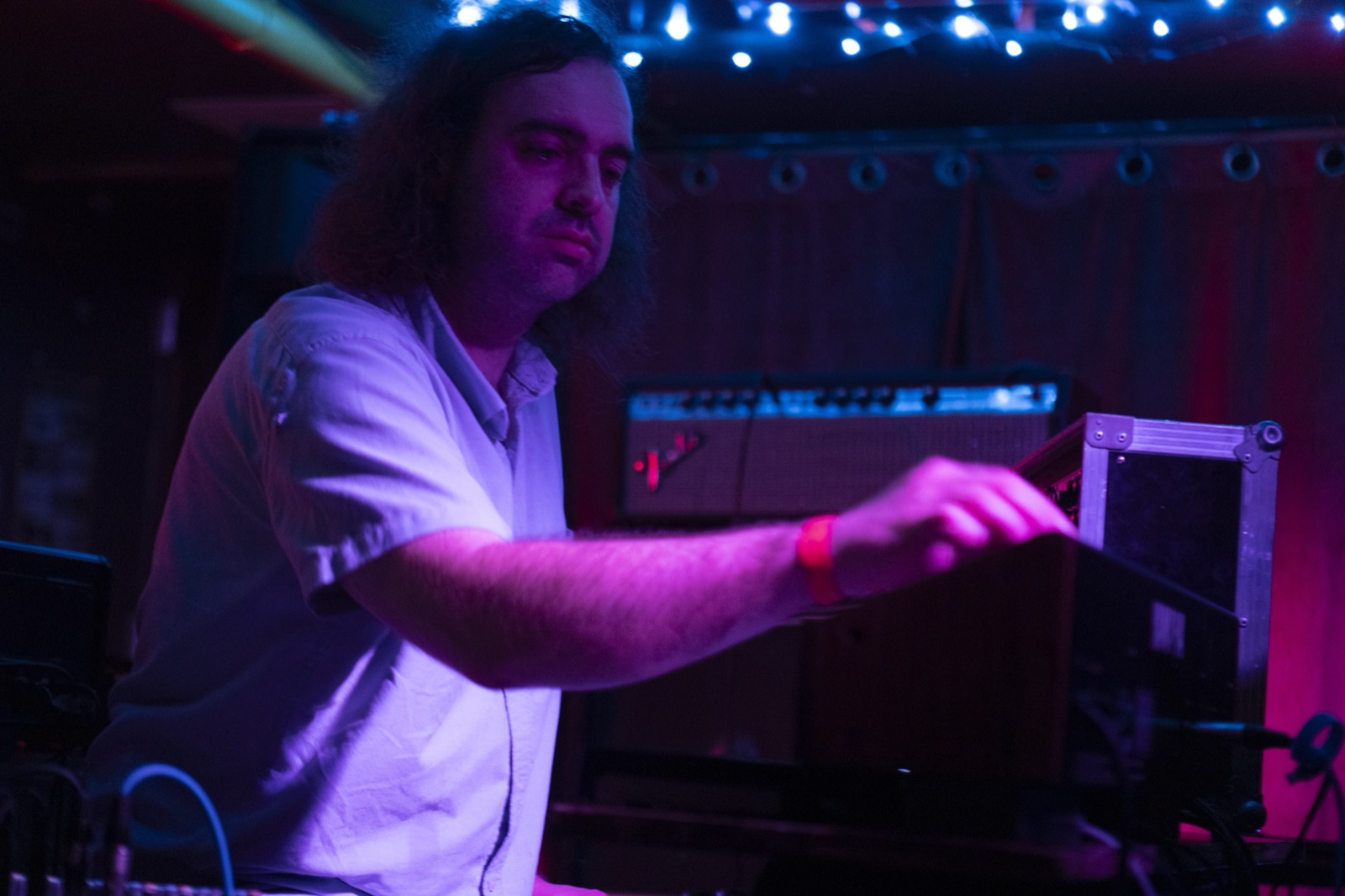 A long-haired keyboardist reaches across his gear in magenta light under a ceiling of blue fairy lights.