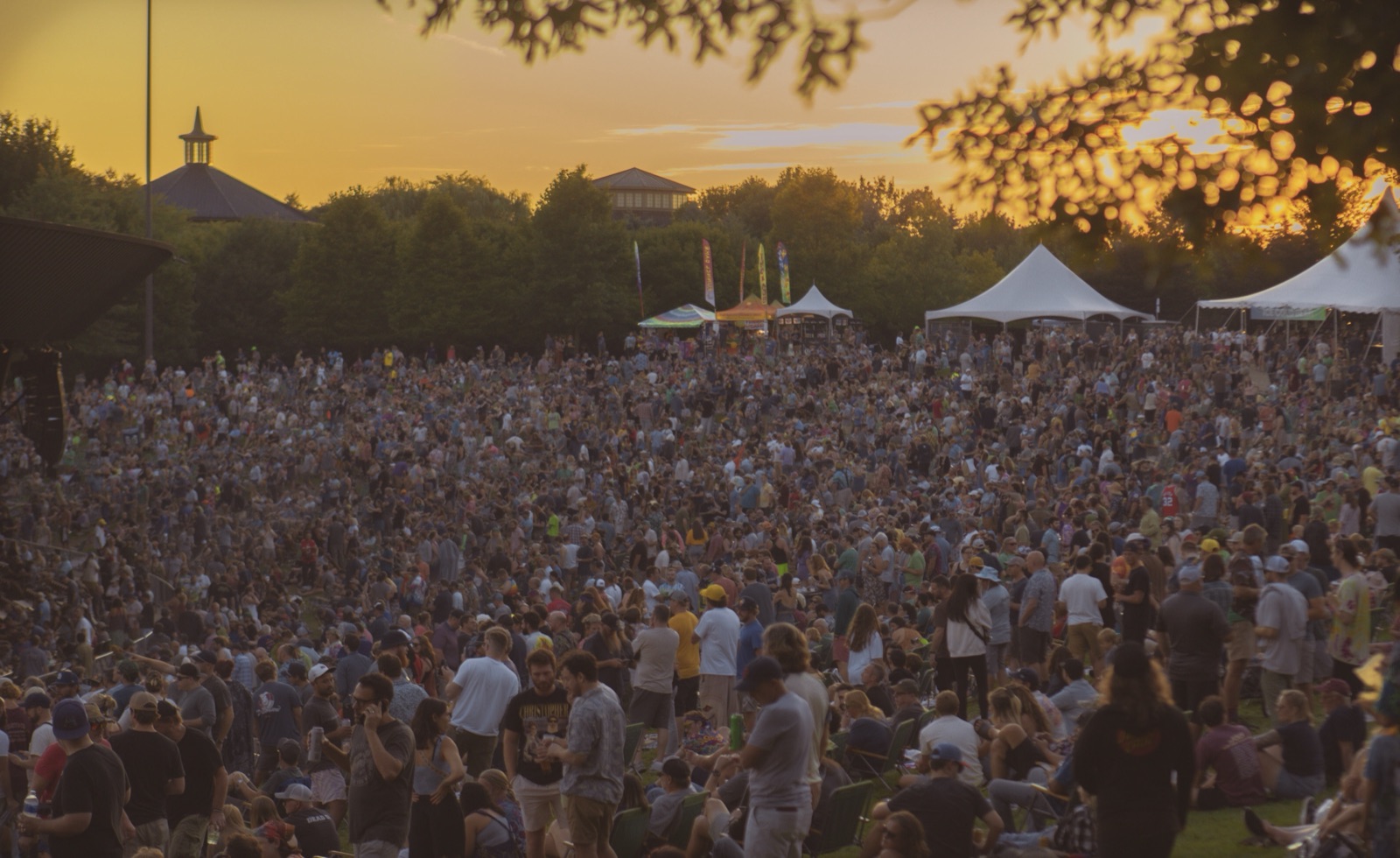 A huge crowd covers the lawn at golden sunset, with white tents and flags behind and leaves framing the top.
