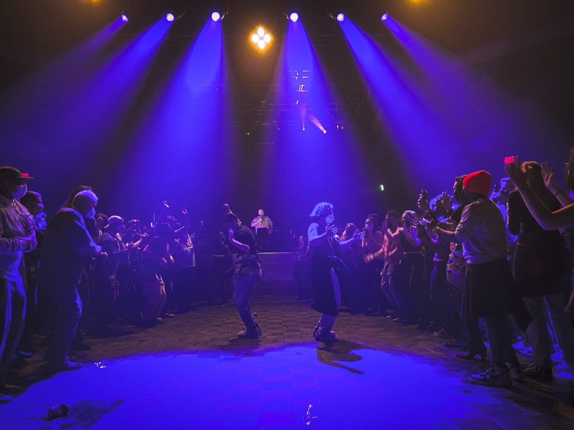 Blue light beams fan down over a ring of dancers on an open floor, with the performer at his table beyond them.