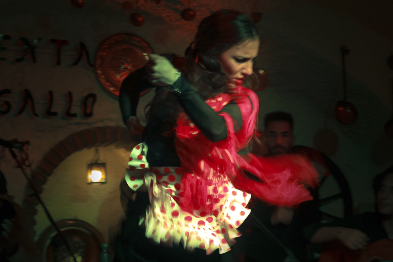A flamenco dancer in a red polka-dot dress spins with her head bowed under red and green light in a whitewashed cave.