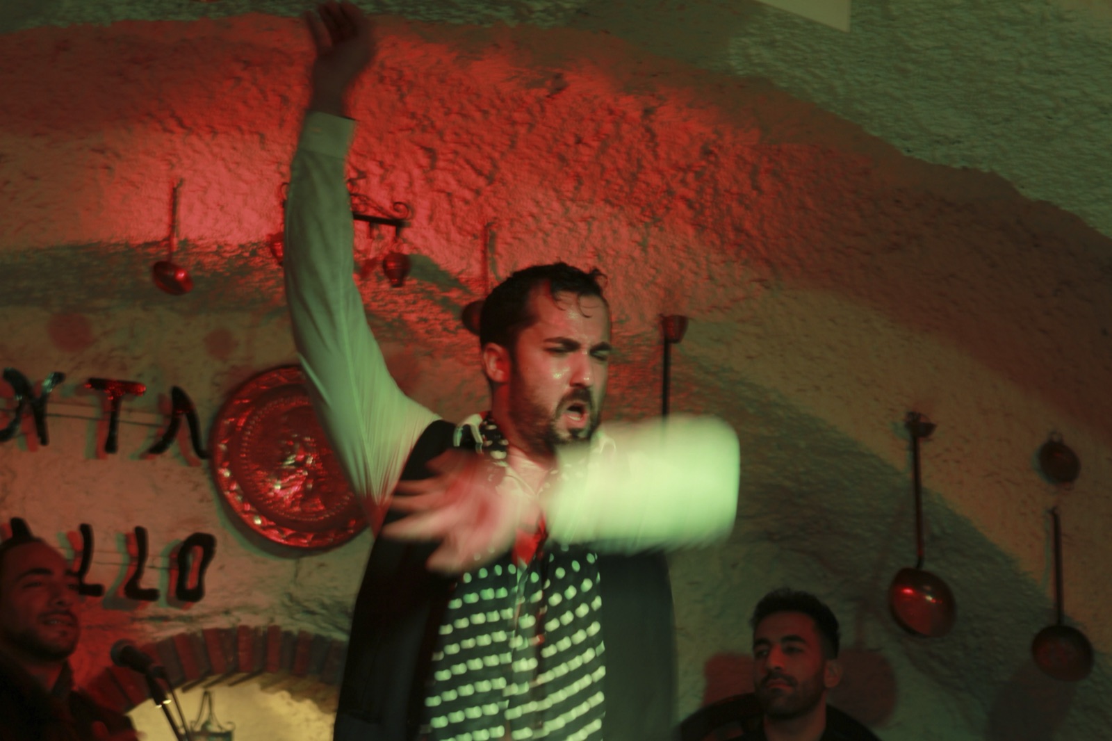 A male flamenco dancer calls out with one arm raised and the other a blur, lit red and green.