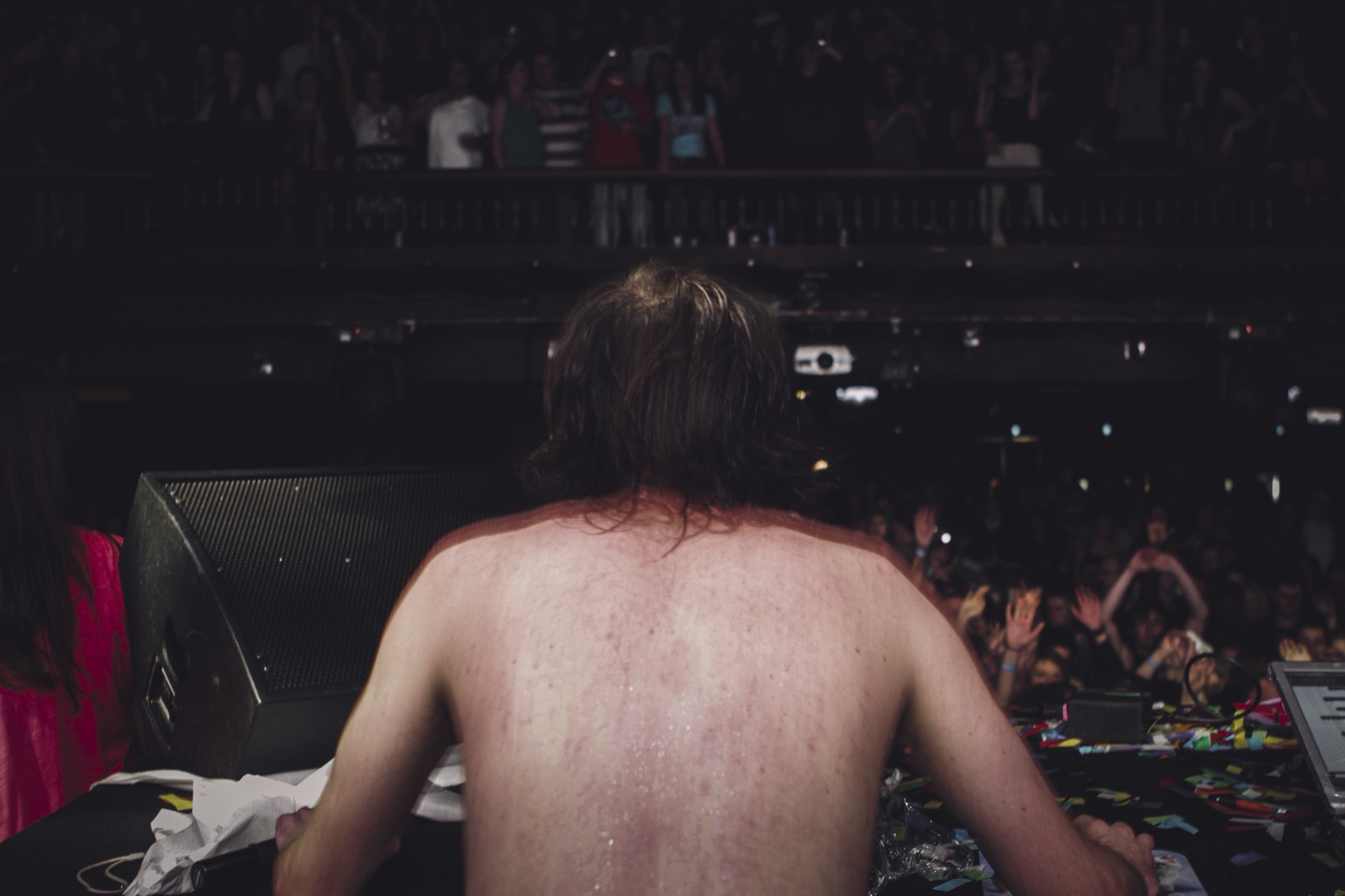 Seen from behind, a shirtless, sweat-soaked Girl Talk faces a packed theater crowd with hands raised and a full balcony above.