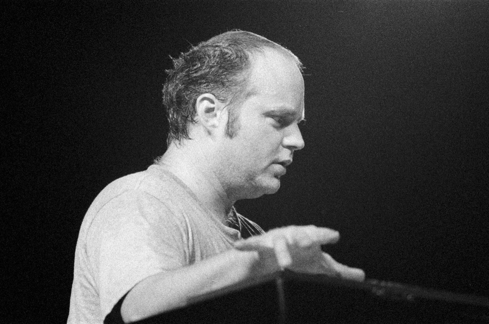 In black and white, the organist plays in profile with his hands raised over the keys against a black background.