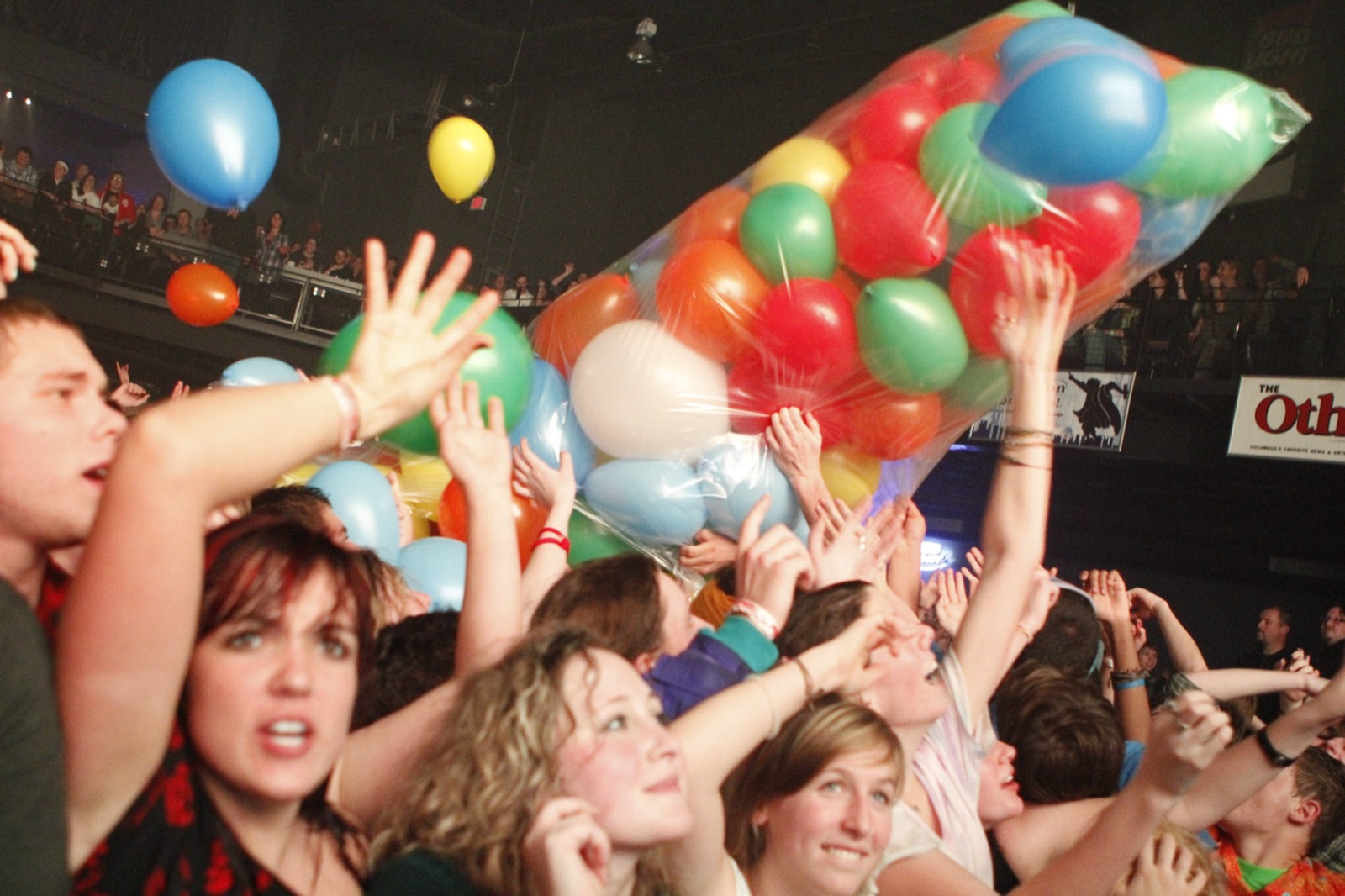 A giant clear bag of balloons floats over a crowd reaching up with open hands.