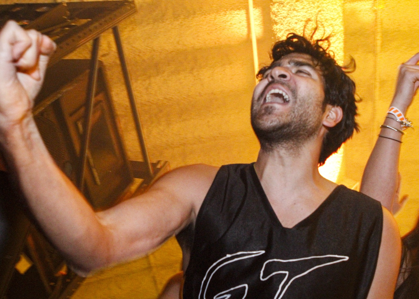 A man in a black jersey shouts with his fist raised in warm yellow light beside a speaker stack on stage.