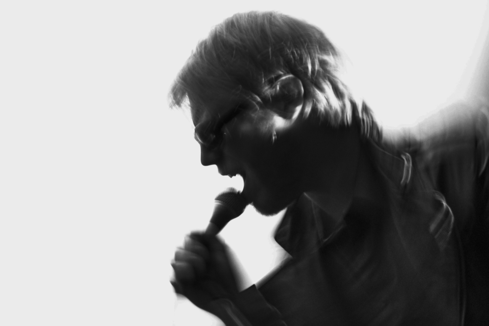A singer shouts into a microphone in dark silhouette against a white wall.