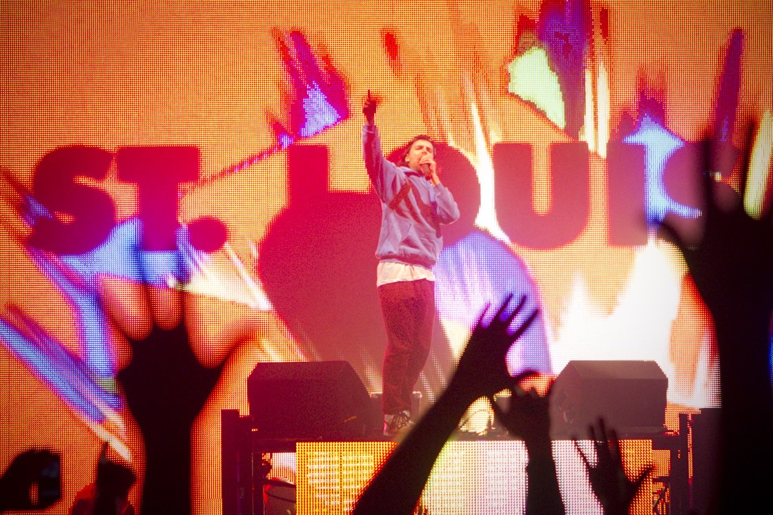 Girl Talk points skyward with a microphone in front of a giant orange and pink ST. LOUIS video wall as fans raise their hands.