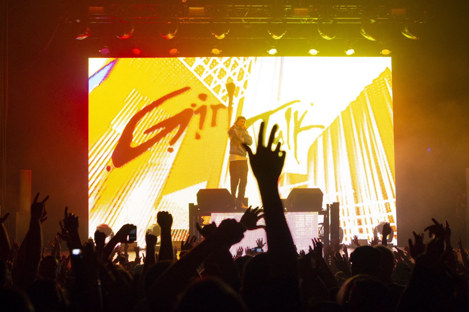 A crowd of raised hands faces a yellow Girl Talk video screen, with the performer standing on the DJ table under the lighting rig.