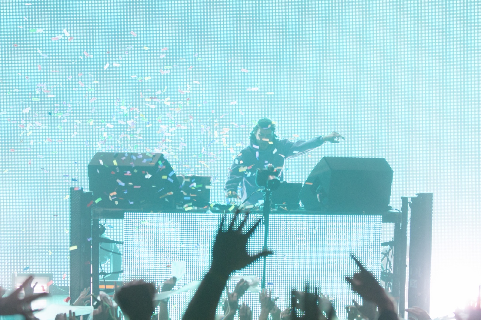 Confetti rains against a cyan screen as the DJ flings an arm out over the booth and the crowd's hands reach up.
