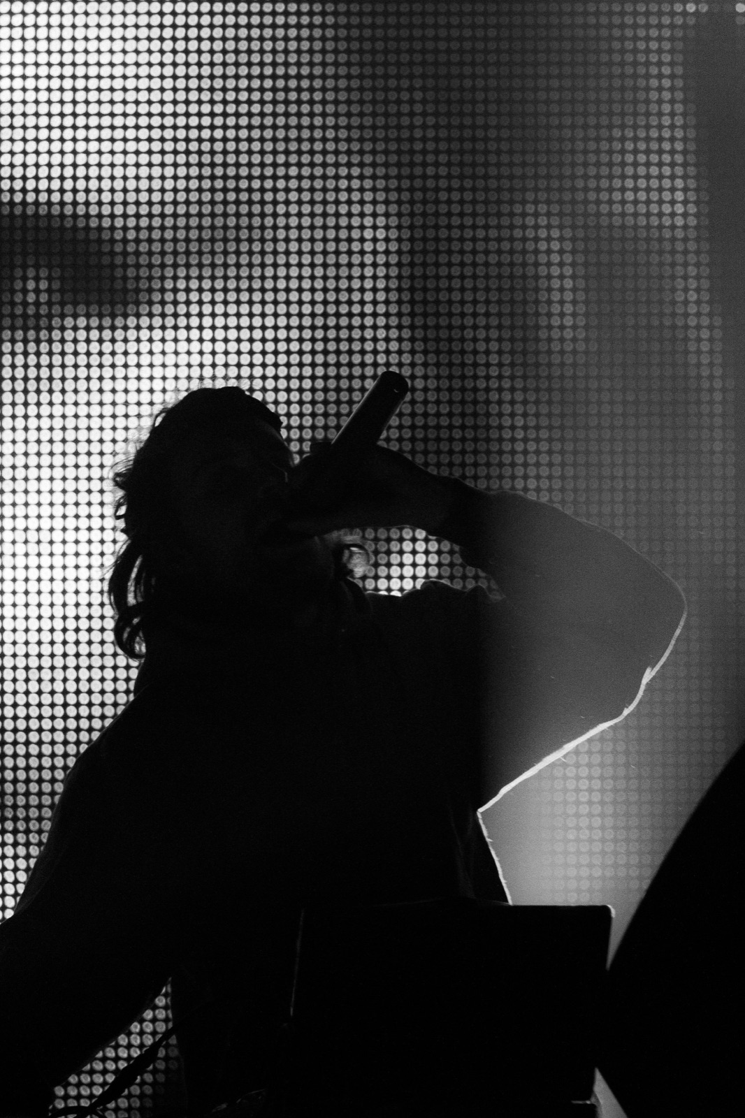 Black and white silhouette of Girl Talk shouting into a microphone in front of a glowing LED dot wall.
